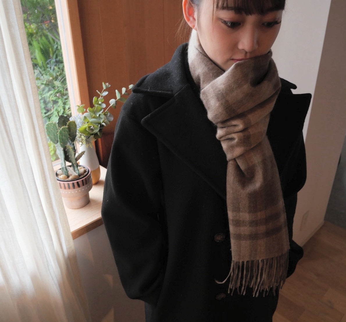 ブラウンチェックのカシミヤストールを黒いコートに合わせた女性の着用例。自然に視線を落とした表情と、フリンジまでの落ち感