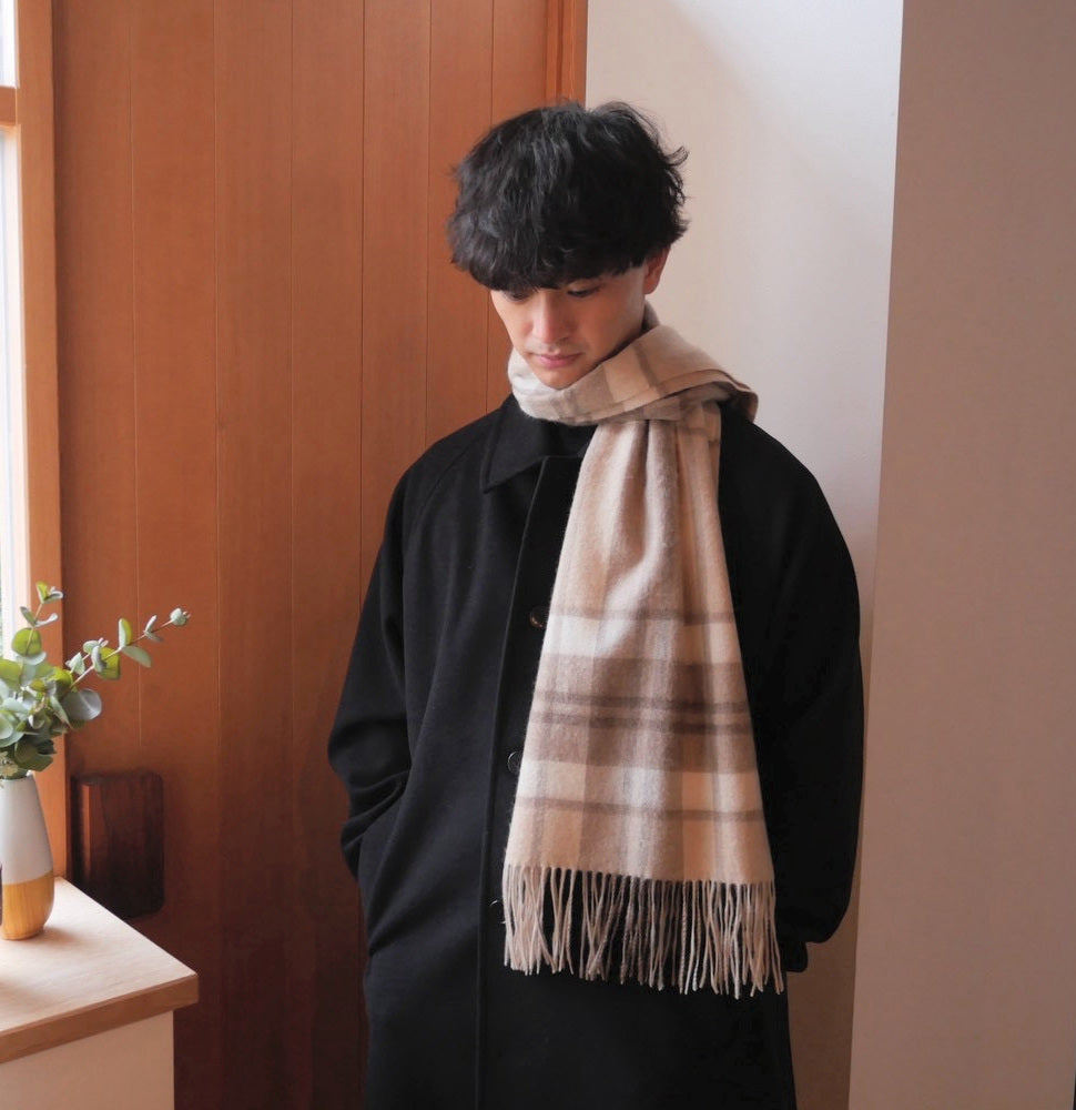 ラインチェックのカシミヤストールを黒いコートに合わせた男性の着用例。手をポケットに入れ、視線を落とした自然な立ち姿とフリンジ