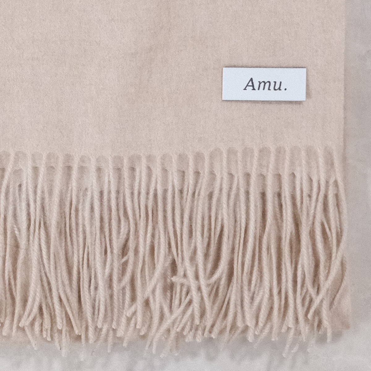 グレージュのカシミヤ生地とフリンジ、Amu.の織りネームの接写