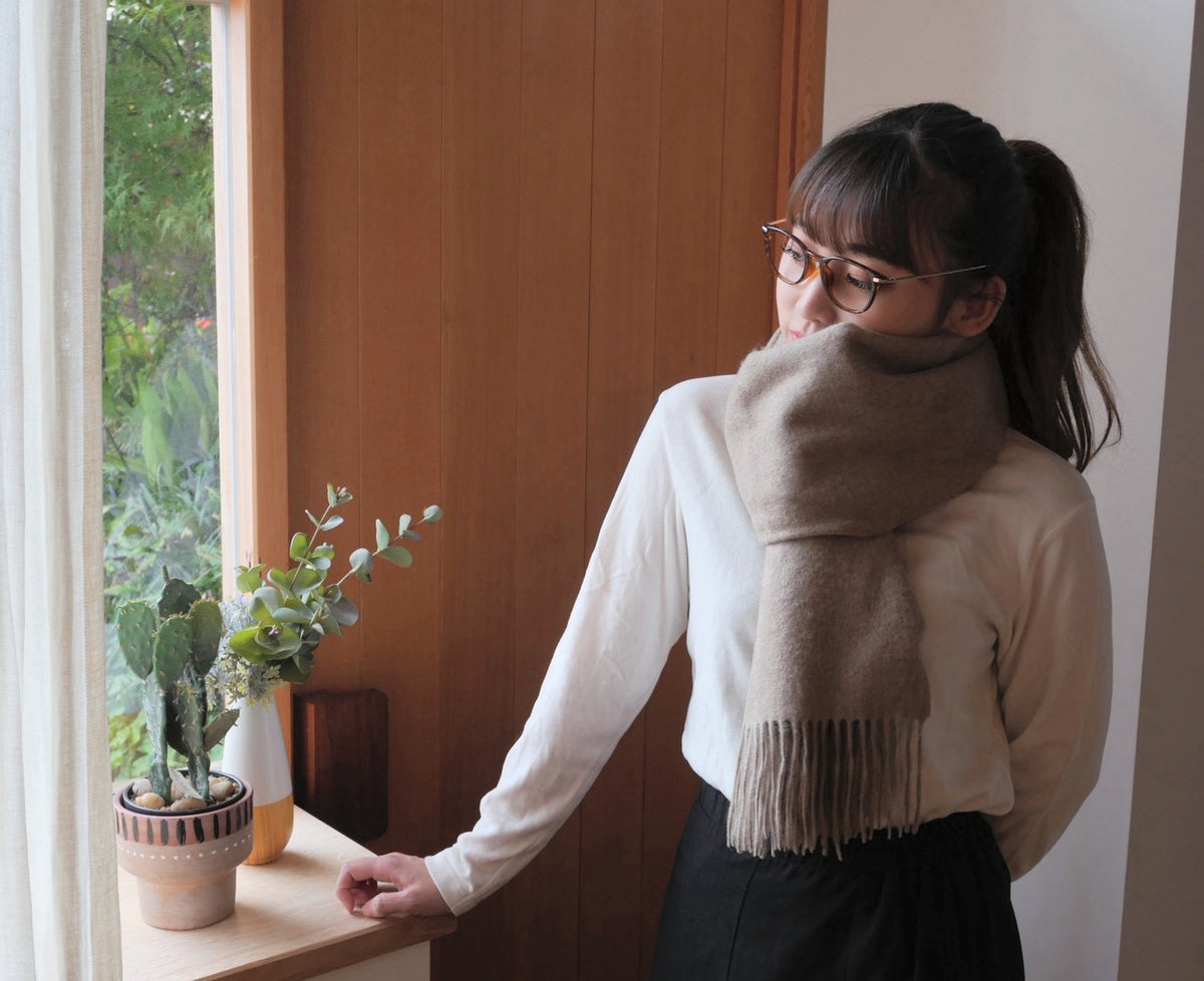 モカブラウンのカシミヤストールを白いニットに合わせ、窓の外へ視線を向けた女性の着用例。巻きのふくらみと全フリンジ