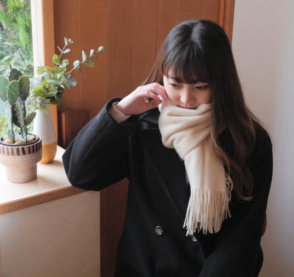 オフホワイトのカシミヤストールを黒いコートに合わせた女性の着用例。窓辺で視線を落とした様子とフリンジ