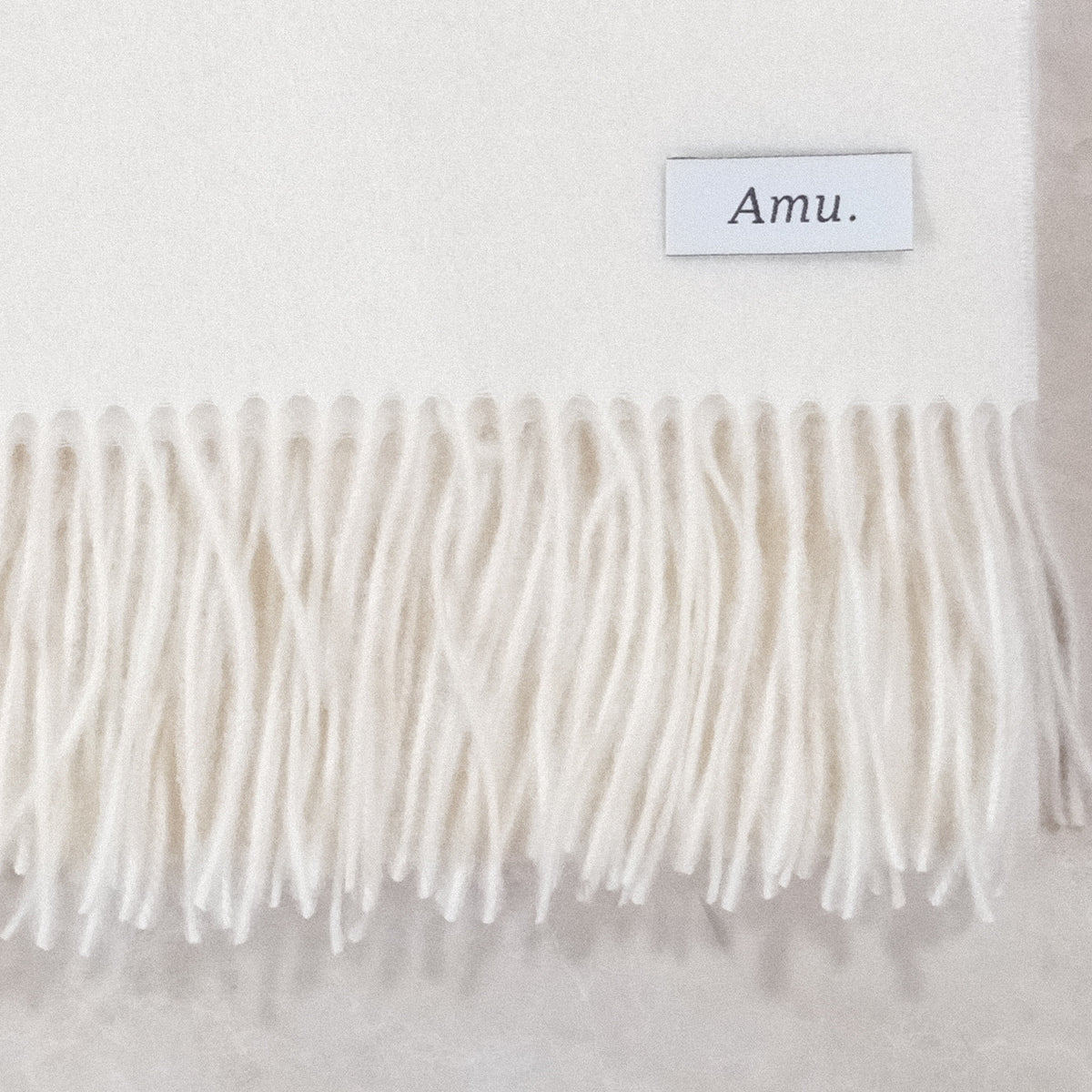 オフホワイトのカシミヤ生地とフリンジ、Amu.の織りネームの接写