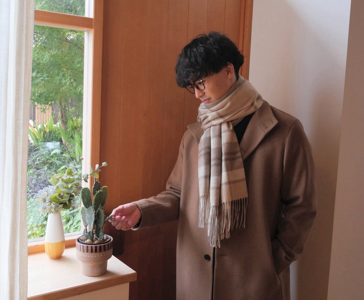 ラインチェックのカシミヤストールをブラウンのコートに合わせ、眼鏡をかけて窓辺に立つ男性の着用例