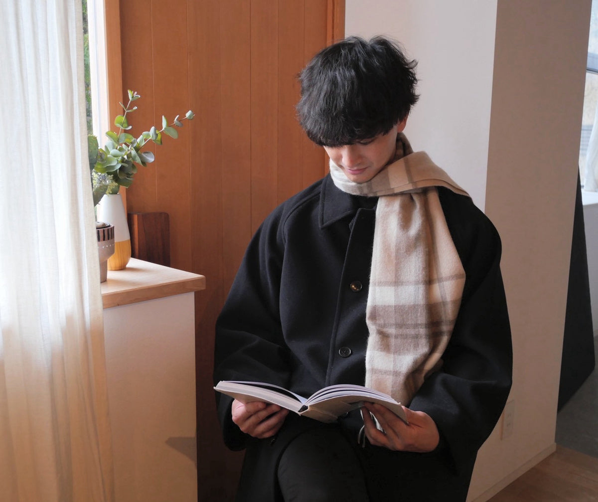 ラインチェックのカシミヤストールをゆるく巻き、黒いコートで本を読む男性。日常の着用イメージ